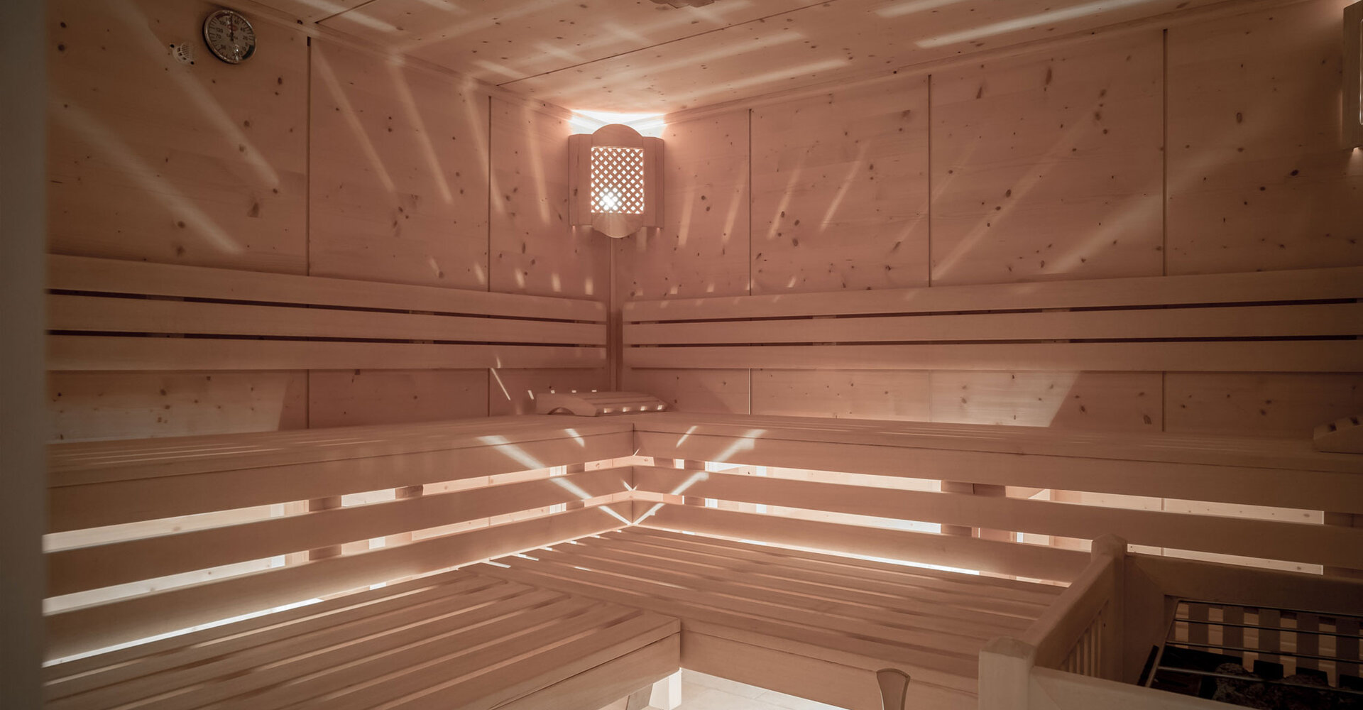 Eine geräumige Holzsauna mit abgestuften Bänken, einem Eimer und einer Kelle, einem Wandofen und sanftem natürlichem Licht, das durch Lamellen strömt und Muster an den Wänden und Bänken erzeugt.