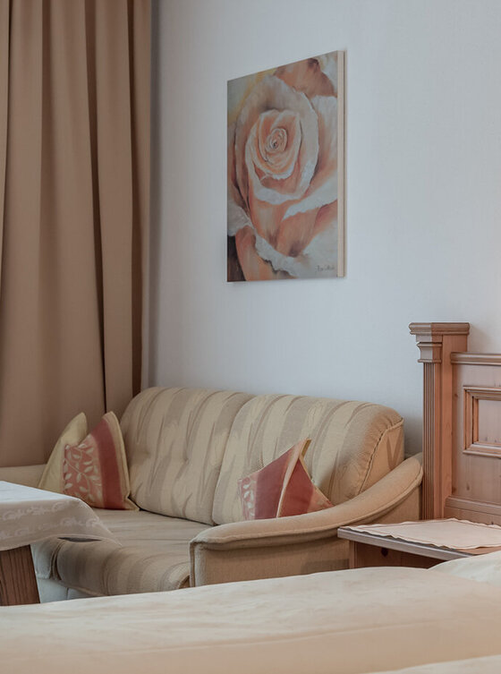 Ein gemütliches Zimmer mit beigen Wänden, einem gepolsterten Sofa, einem kleinen Tisch mit Tischtuch, zwei Betten mit cremefarbener Bettwäsche und einem Rosengemälde an der Wand über dem Sofa. Die warme Beleuchtung schafft eine gemütliche Atmosphäre.