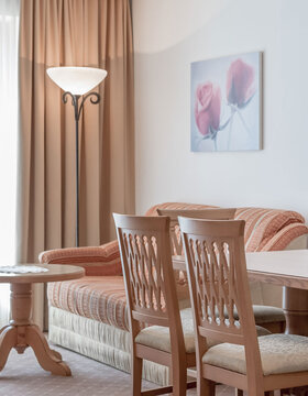 Ein gemütliches Wohnzimmer mit einem pfirsichfarbenen Sofa, Holzstühlen an einem Tisch, einem runden Beistelltisch, einer hohen Stehlampe, beigen Vorhängen und einem Wandbild mit zwei rosa Blumen.