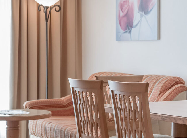 Ein gemütliches Wohnzimmer mit einem pfirsichfarbenen Sofa, Holzstühlen an einem Tisch, einem runden Beistelltisch, einer hohen Stehlampe, beigen Vorhängen und einem Wandbild mit zwei rosa Blumen.