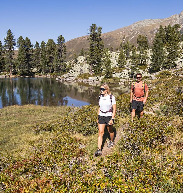 Zwei Personen wandern auf einem Pfad an einem ruhigen See, der von Bäumen und Büschen umgeben ist, mit Bergen im Hintergrund unter einem klaren blauen Himmel.