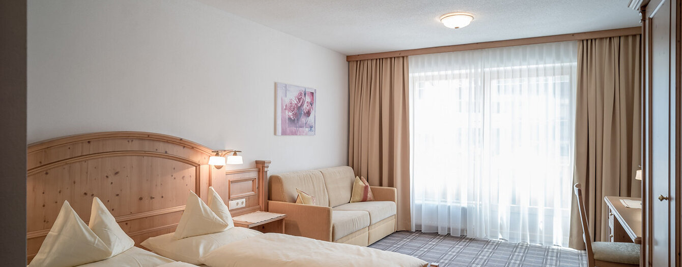 Ein helles Apartmentzimmer mit einem Doppelbett, einem hellen Sofa, Holzmöbeln, einem Schreibtisch und großen Fenstern mit durchsichtigen Vorhängen, die das Tageslicht hereinlassen.