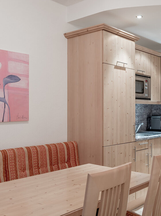 Moderne Küche mit hellen Holzschränken, eingebauter Mikrowelle, Geschirrspüler, Kaffeemaschine und grauer Aufkantung. Im Vordergrund ein Esstisch aus Holz mit Stühlen und einer gepolsterten Bank. An der Wand hängt ein Blumengemälde.