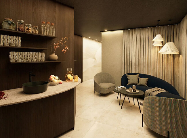 Eine gemütliche Wellness Lounge mit einem geschwungenen Sofa, zwei Sesseln, einem runden Couchtisch und Pendelleuchten. Regale mit Gläsern und Dekoration, eine Arbeitsplatte mit Obst und helle Vorhänge vervollständigen den warmen, einladenden Raum.