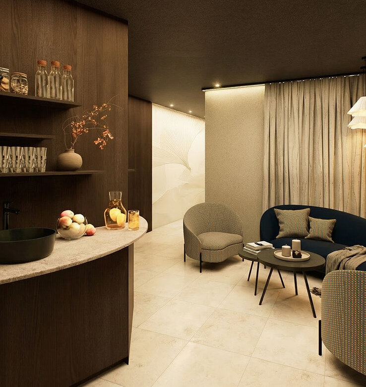 Eine gemütliche Wellness Lounge mit einem geschwungenen Sofa, zwei Sesseln, einem runden Couchtisch und Pendelleuchten. Regale mit Gläsern und Dekoration, eine Arbeitsplatte mit Obst und helle Vorhänge vervollständigen den warmen, einladenden Raum.