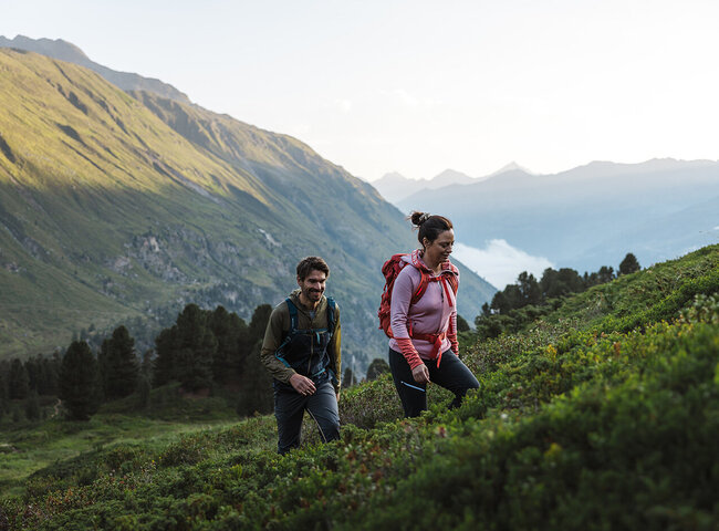 Zwei Personen mit Rucksäcken wandern auf einem grünen Berghang mit malerischen Hügeln und Bäumen im Hintergrund.