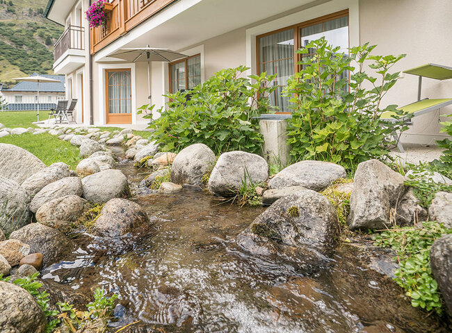 Ein kleiner Bach fließt über Felsen neben einem Gebäude mit Balkonen, die mit bunten Blumen und hölzernen Geländern geschmückt sind, vor grünen Hügeln unter einem teilweise bewölkten Himmel.