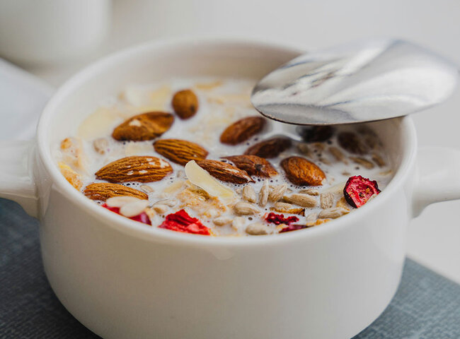 Eine weiße Schüssel mit Müsli, Milch, ganzen Mandeln und Trockenobststücken auf einem grauen Tischset, in der ein Metalllöffel liegt.