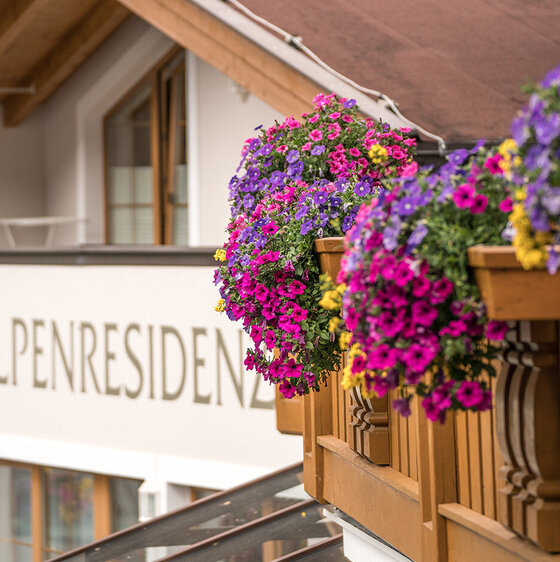 Holzbalkon mit bunten Blumen in Hängekörben, der an ein Gebäude im alpinen Stil angebaut ist. Das Wort ALPENRESIDENZ ist auf der weißen Wand des Gebäudes im Hintergrund zu sehen.