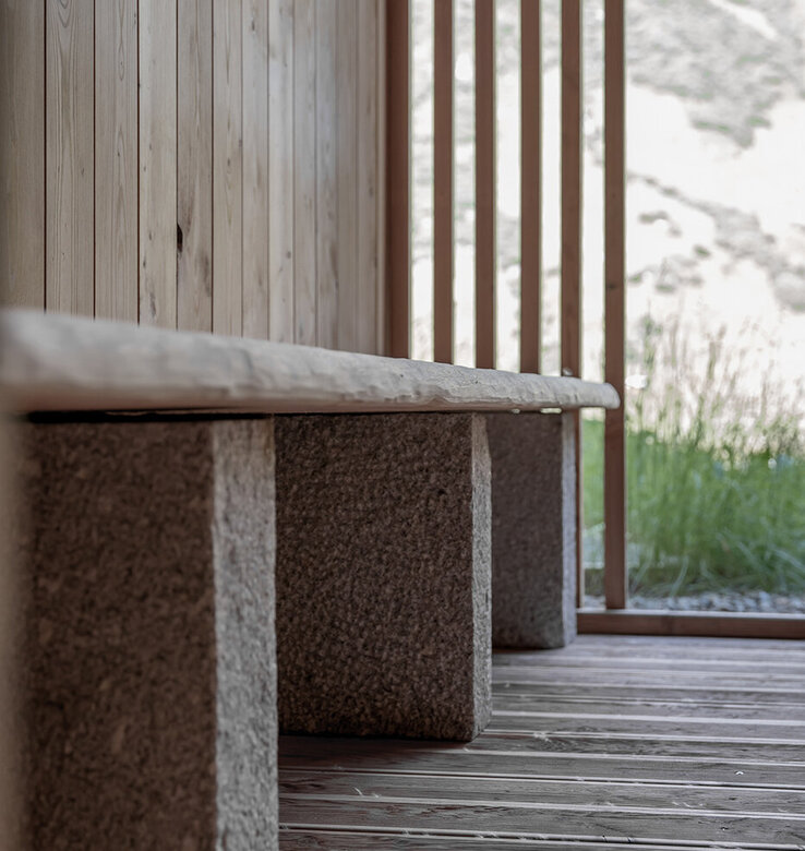 Eine minimalistische Holzbank mit steinernen Stützen steht auf einem Holzboden, neben vertikalen Holzlatten und einer Wand, während draußen Grün und natürliches Licht zu sehen sind.