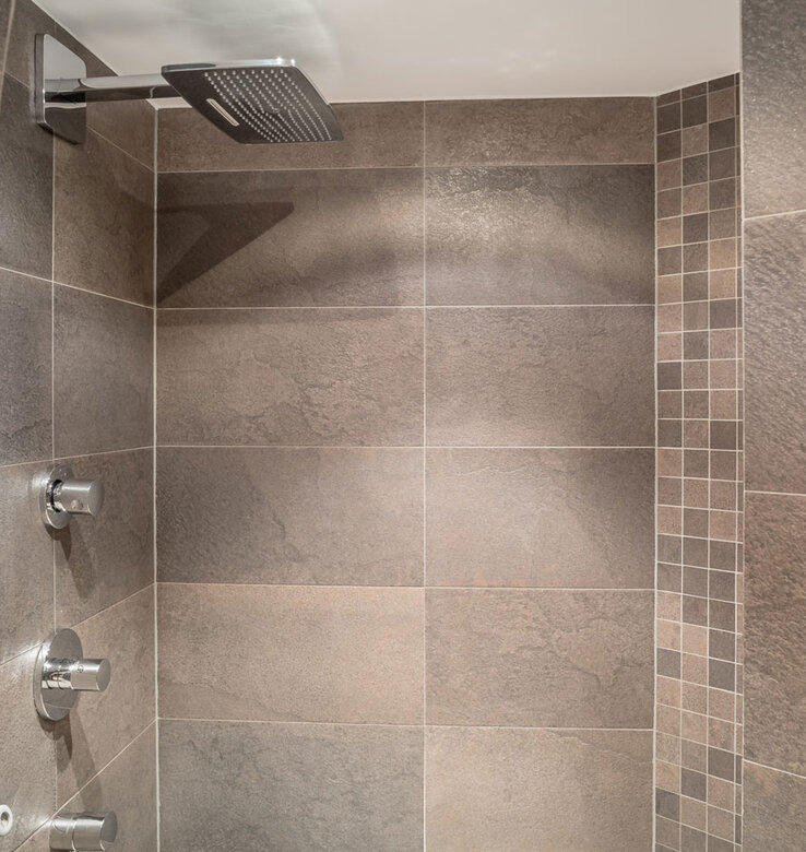 Moderne Dusche mit großer, quadratischer Regendusche und zwei Bedienknöpfen. Die Wände sind mit braunen und grauen Fliesen bedeckt, mit einem vertikalen Streifen aus kleinen Mosaikfliesen auf der rechten Seite.