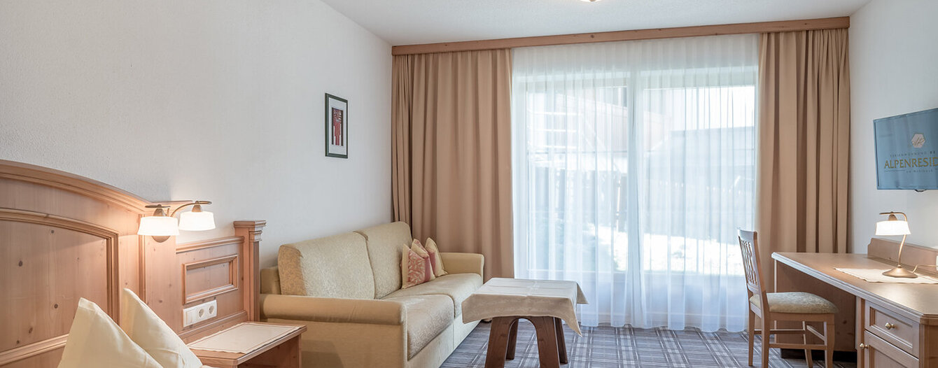 Ein helles Apartmentzimmer mit einem Bett, einem hölzernen Kopfteil, einem beigen Sofa, einem Couchtisch, einem Schreibtisch, einem Stuhl und einem großen Fenster mit durchsichtigen Vorhängen, die das natürliche Licht hereinlassen.