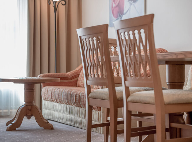 Ein gemütliches Wohnzimmer mit Holzstühlen an einem Esstisch, einem runden Couchtisch aus Holz und einem gemusterten Sofa neben einem hellen Fenster mit Vorhängen.