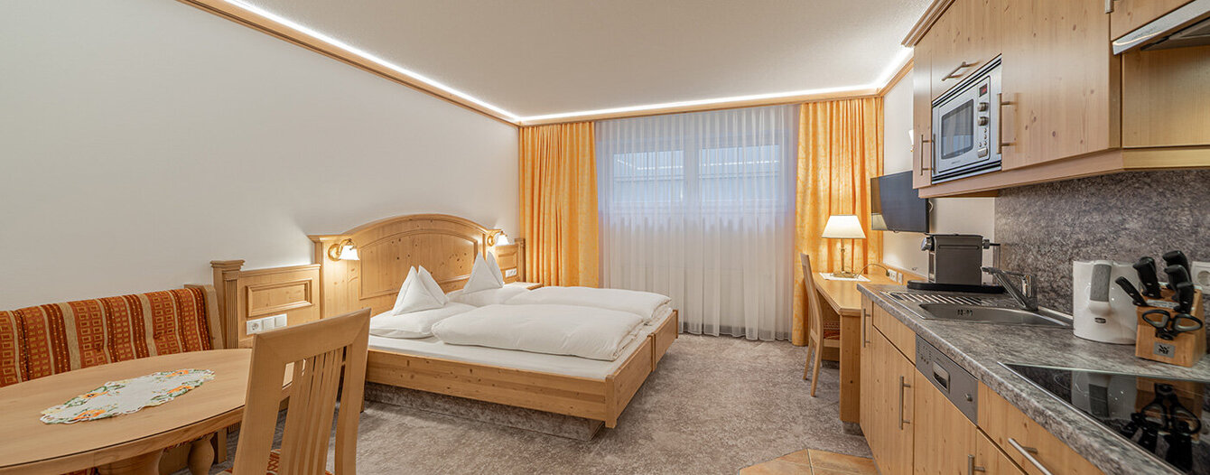Ein gemütliches Hotelzimmer mit einem Doppelbett, einem kleinen runden Esstisch mit zwei Stühlen, einer eingebauten Küchenzeile aus Holz und einem großen Fenster mit durchsichtigen Vorhängen und orangefarbenen Gardinen. Die warme, stimmungsvolle Beleuchtung schafft eine einladende Atmosphäre.