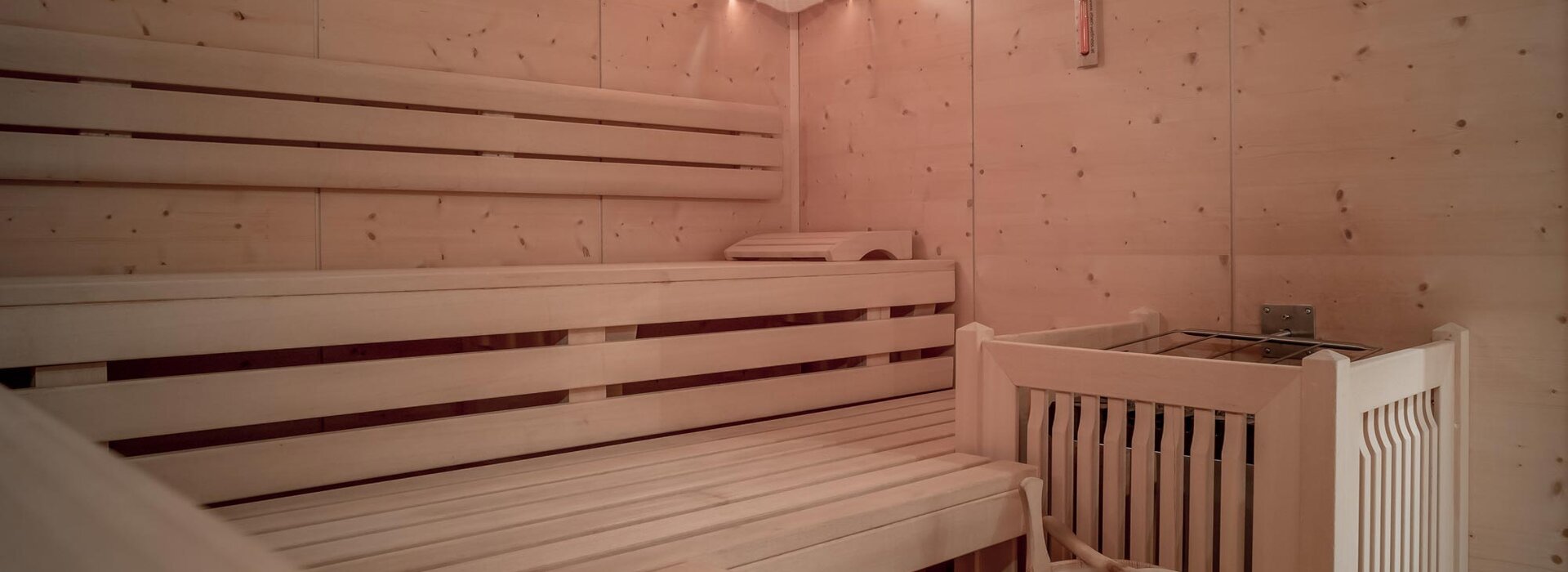 Eine saubere, leere Holzsauna mit Stufenbänken, einem Ofen mit Steinen in einer Ecke, einem Holzeimer mit Schöpfkelle und warmer Beleuchtung an den kiefergetäfelten Wänden.