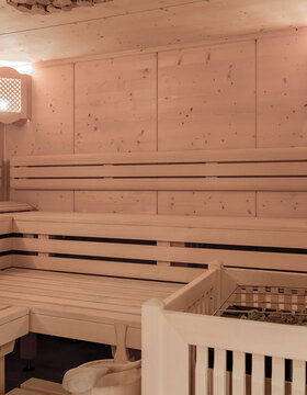 Eine geräumige, helle Holzsauna mit gestuften Bänken an den Wänden, sanfter Beleuchtung in den Ecken und einem Ofen mit Steinen im Vordergrund.
