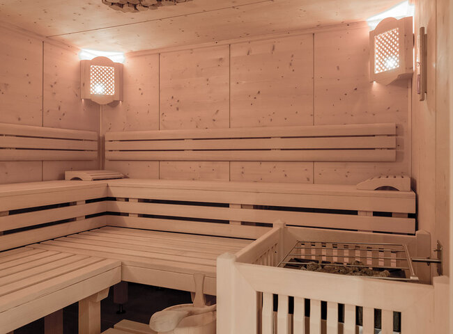 Eine geräumige, helle Holzsauna mit gestuften Bänken an den Wänden, sanfter Beleuchtung in den Ecken und einem Ofen mit Steinen im Vordergrund.