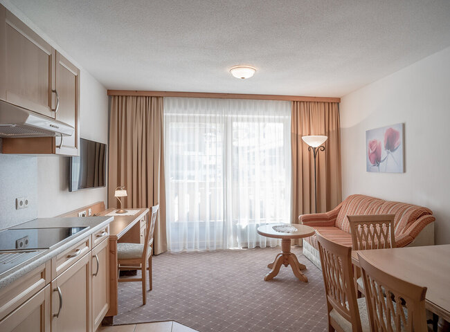 Ein helles, aufgeräumtes Wohnzimmer mit einer Küchenzeile, Holzmöbeln, einem Sofa, einem kleinen runden Tisch, einem Flachbildfernseher und großen, mit Vorhängen versehenen Fenstern, die natürliches Licht hereinlassen. An der weißen Wand hängt ein Blumengemälde.