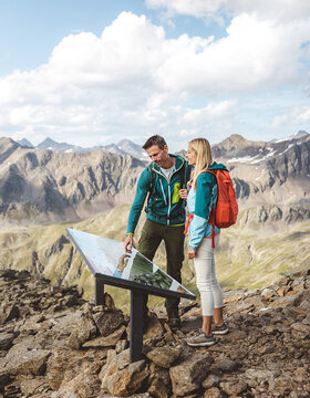 Zwei Wanderer lesen eine Informationstafel auf einem felsigen Bergpfad mit Gipfeln und Wolken im Hintergrund.