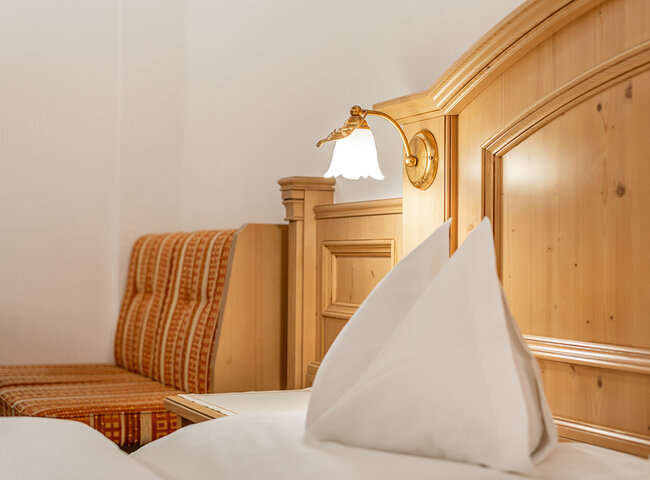 Nahaufnahme eines ordentlich gemachten Bettes mit weißen Kissen und Bettwäsche, daneben ein hölzernes Kopfteil und ein Nachttisch. Eine Wandlampe und eine gepolsterte Bank mit orangefarbenem Stoff sind ebenfalls zu sehen.