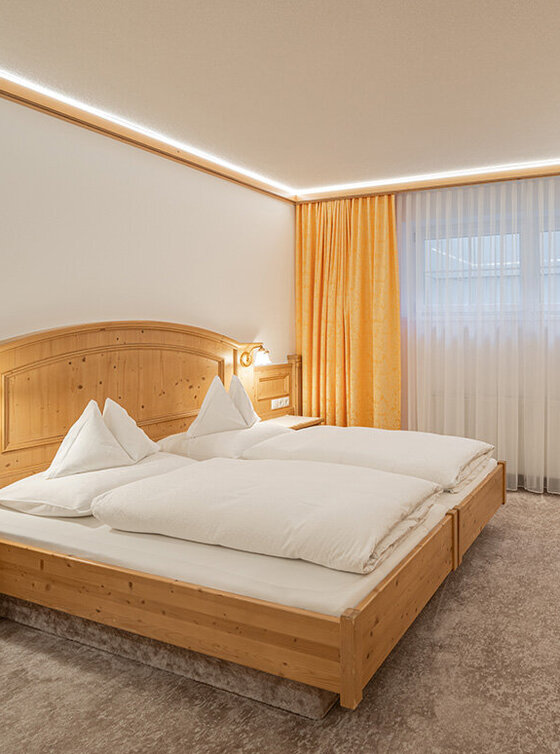 Ein gemütliches Apartmentzimmer mit Doppelbett, Holzmöbeln, Schreibtisch mit Lampe, Flachbildfernseher, Kochnische, Kaffeemaschine und großem Fenster mit durchsichtigen und gelben Vorhängen. Die Beleuchtung ist warm und einladend.