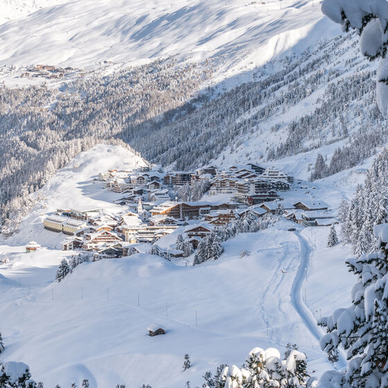 Ein verschneites Alpendorf in einem Tal, umgeben von schneebedeckten Bäumen und Bergen unter einem klaren blauen Himmel. Inmitten der Winterlandschaft sind Skispuren und Dächer zu sehen.