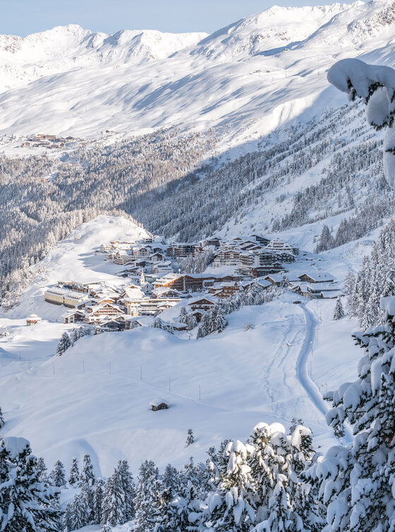 Ein verschneites Alpendorf in einem Tal, umgeben von schneebedeckten Bäumen und Bergen unter einem klaren blauen Himmel. Inmitten der Winterlandschaft sind Skispuren und Dächer zu sehen.