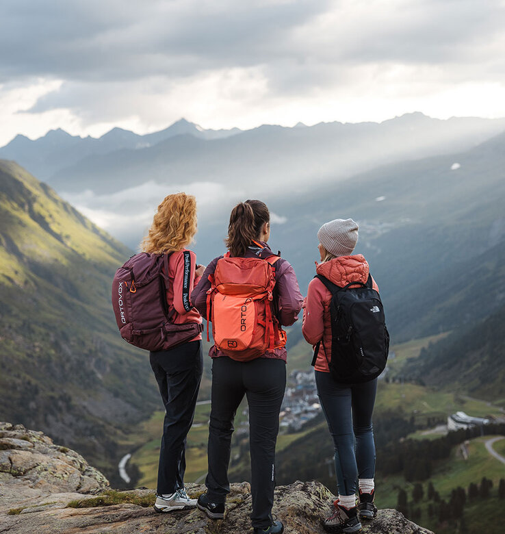 Drei Personen mit Rucksäcken stehen auf einem Felsvorsprung mit Blick auf ein malerisches Bergtal, während das Sonnenlicht durch die Wolken auf die Landschaft darunter fällt.