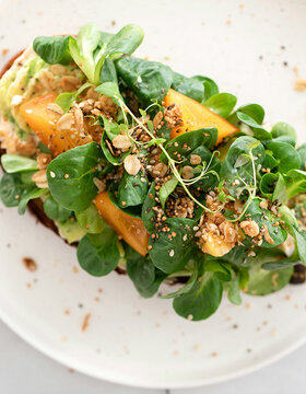 Offenes Sandwich mit Blattgemüse, gelben Tomatenscheiben und gestreuten Samen auf einem weißen Teller, von oben auf einer hellen Fläche fotografiert.