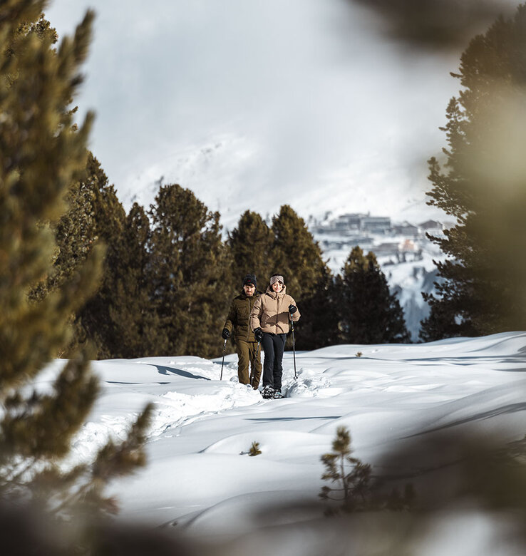 Zwei Personen gehen mit Stöcken durch einen verschneiten Waldweg, umgeben von immergrünen Bäumen, mit Bergen und einem Dorf im Hintergrund.
