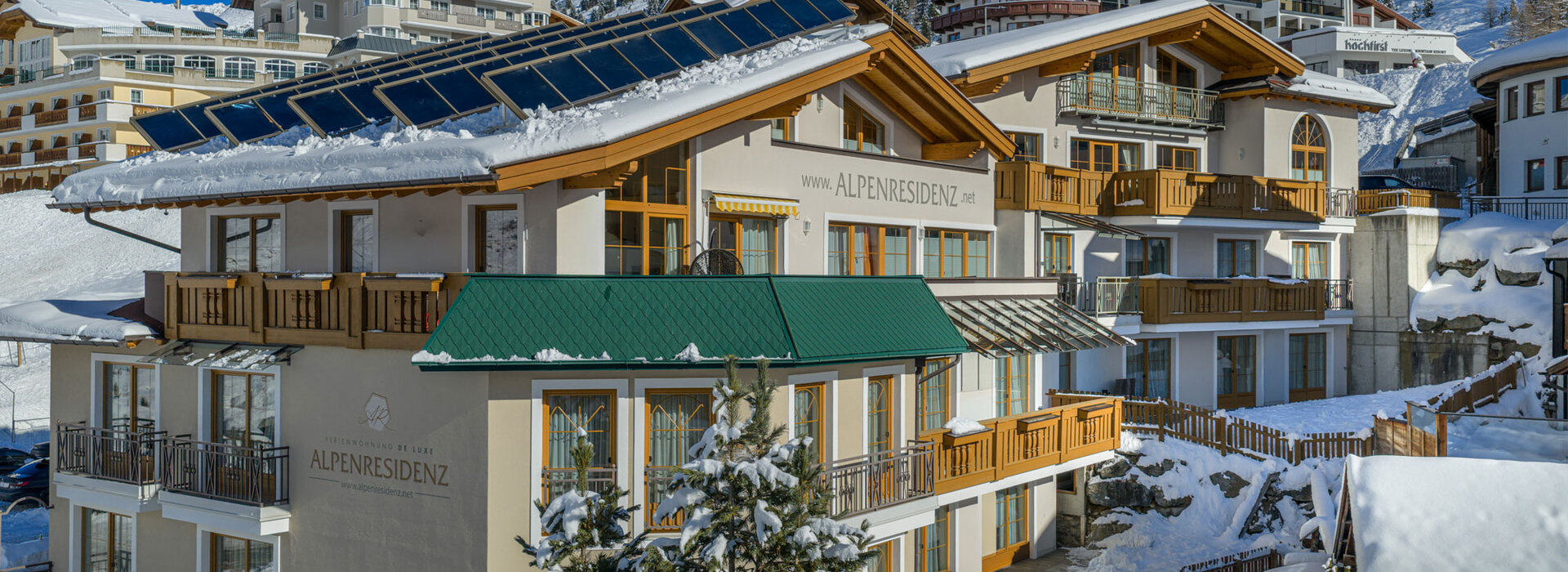 Modern alpines Hotelgebäude mit grünen und hölzernen Akzenten liegen zwischen schneebedeckten Bäumen und Hügeln unter einem klaren blauen Himmel, mit Photovoltaik auf dem Dach und der Berglandschaft im Hintergrund.