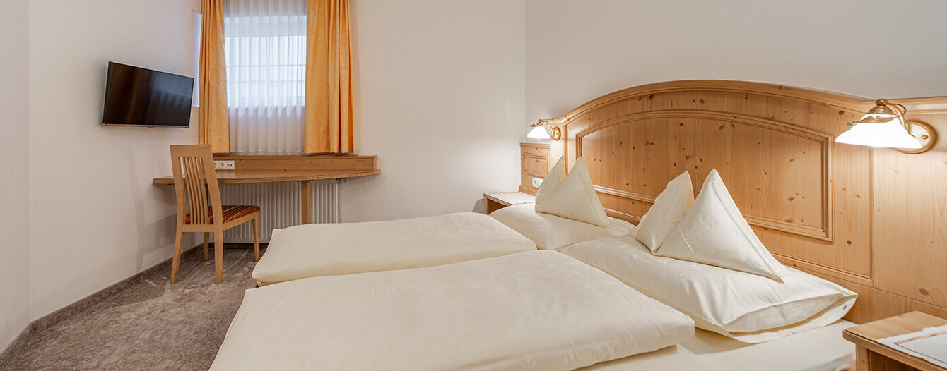 Das gemütliche Apartmentzimmer verfügt über ein Bett mit weißem Bettzeug, ein Kopfteil aus Holz, einen Schreibtisch mit Stuhl, einen an der Wand montierten Fernseher und ein Fenster mit gelben Vorhängen. Das Zimmer hat neutrale Wände und Teppichböden.