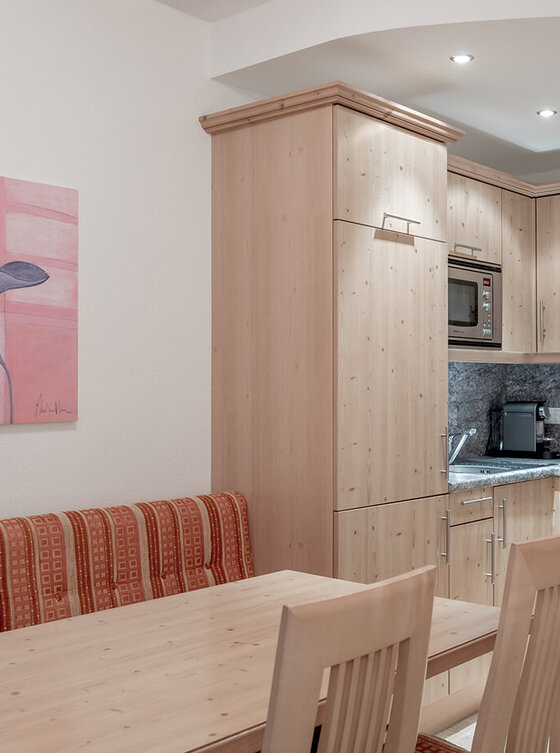Eine kompakte Küche mit hellen Holzschränken, Einbaugeräten und einer grauen Aufkantung. Ein hölzerner Esstisch mit zwei Stühlen und einer gepolsterten Bank steht in der Nähe, und an der Wand hängt ein rosa Blumengemälde.