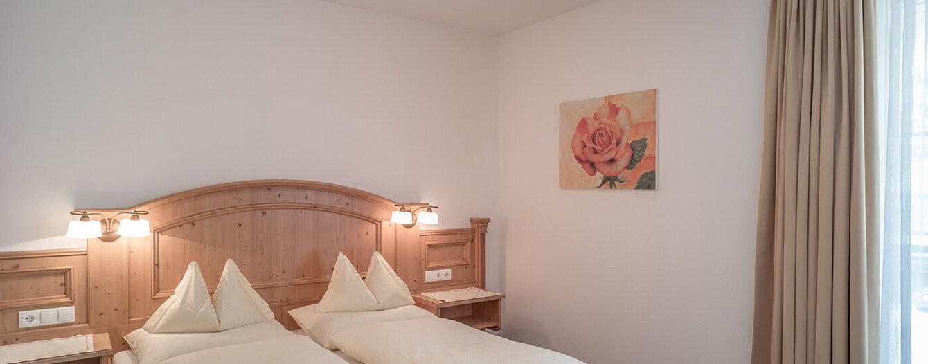 Ein gemütliches Apartmentzimmer mit einem Doppelbett, hellem Holzkopfteil, zwei Kissen, Nachttischen mit Lampen, kariertem Teppich, beigen Vorhängen und einem Wandbild mit einer rosa Rose. Sanfte Beleuchtung und minimalistisches Dekor.