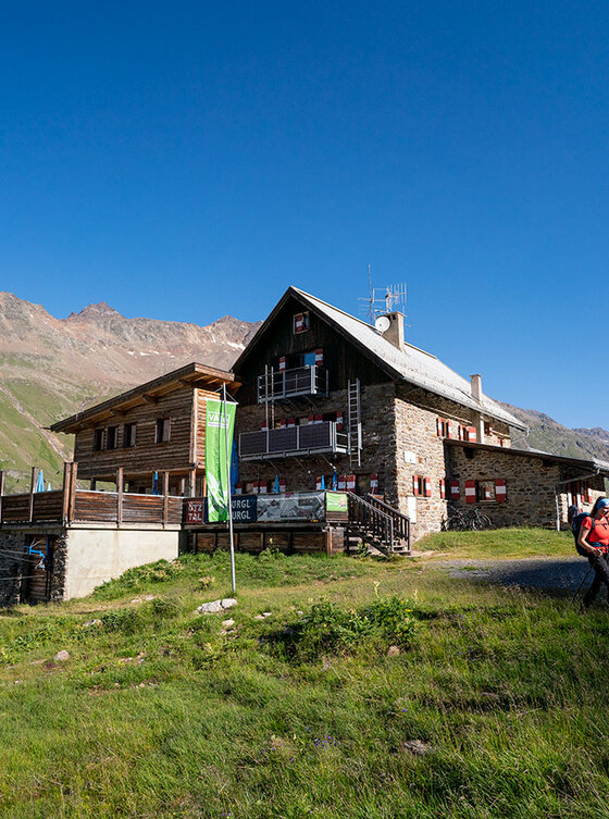 Eine Berghütte mit einer Holzterrasse und Menschen, die auf einem grasbewachsenen Weg unter einem klaren blauen Himmel spazieren gehen.