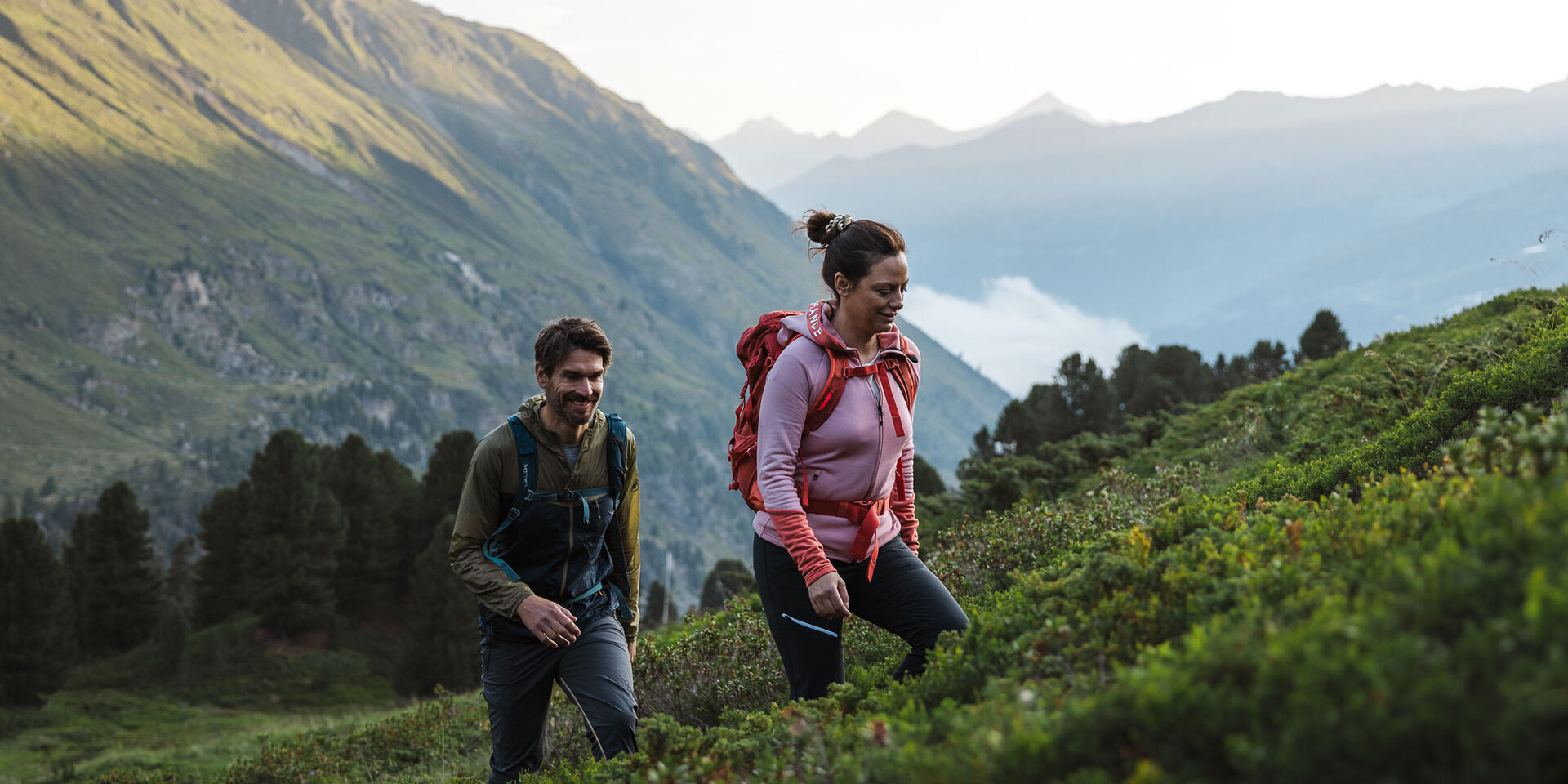 Ein Mann und eine Frau wandern mit Rucksäcken einen grasbewachsenen, grünen Berghang hinauf, umgeben von Bäumen und entfernten Berggipfeln unter einem teilweise bewölkten Himmel.