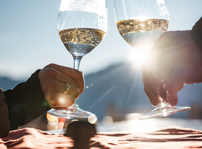 Zwei Personen stoßen im Freien im Sonnenlicht mit Weingläsern an, mit einem unscharfen landschaftlichen Hintergrund und einem Tisch im Vordergrund.