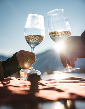 Zwei Personen stoßen im Freien im Sonnenlicht mit Weingläsern an, mit einem unscharfen landschaftlichen Hintergrund und einem Tisch im Vordergrund.