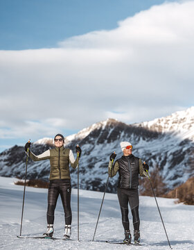 Zwei Personen stehen mit Langlaufski auf verschneitem Boden, gekleidet in Winterkleidung, mit Bergen und blauem Himmel im Hintergrund.