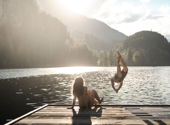 Eine Person sitzt auf einem hölzernen Steg an einem See, während eine andere Person ins Wasser springt, umgeben von bewaldeten Hügeln und Sonnenlicht, das durch die Bäume fällt.