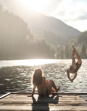 Eine Person sitzt auf einem hölzernen Steg an einem See, während eine andere Person ins Wasser springt, umgeben von bewaldeten Hügeln und Sonnenlicht, das durch die Bäume fällt.