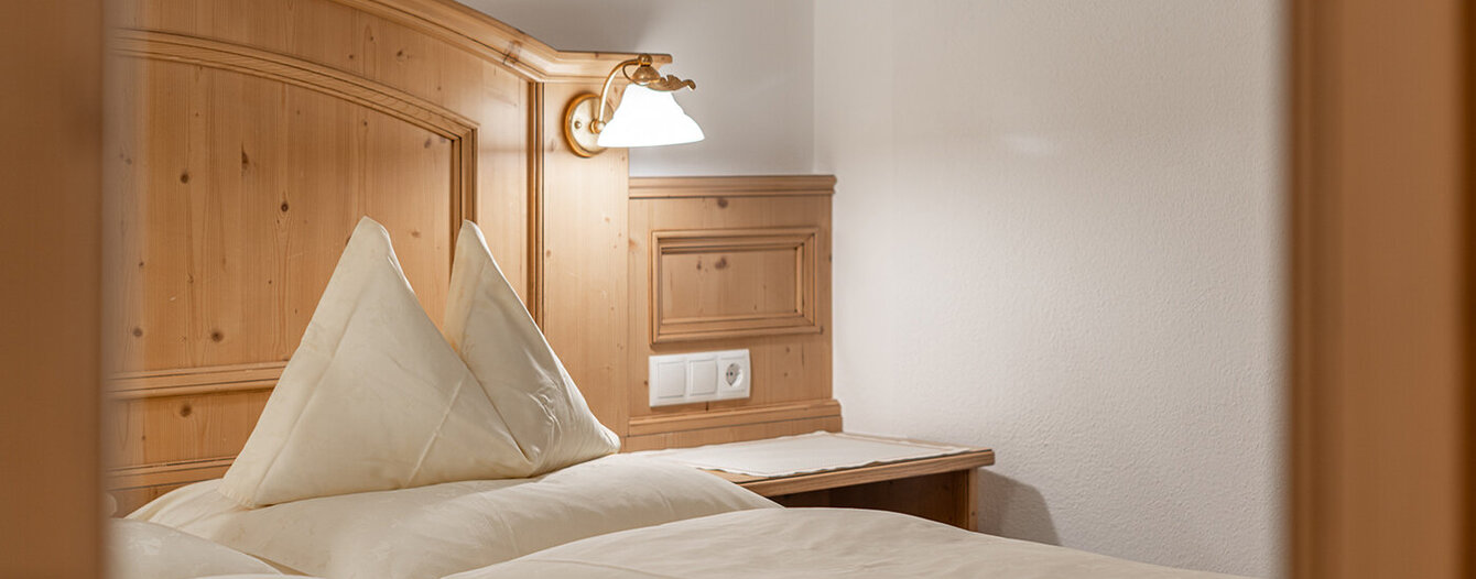 Ein gemütliches Schlafzimmer mit hellen Holzmöbeln, einem Bett mit cremefarbenem Bettzeug und einer Wandlampe über einem hölzernen Kopfteil und einem kleinen Schreibtisch.