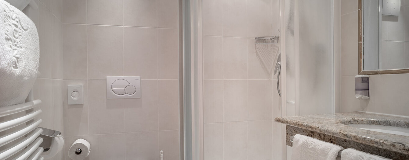 Modernes Badezimmer mit Glasdusche, wandmontierter Toilette, Handtuchhalter mit gestapelten weißen Handtüchern, Toilettenpapierhalter und einem Waschbecken mit Marmorarbeitsplatte und gefalteten Handtüchern. Die Einrichtung ist sauber und minimalistisch.