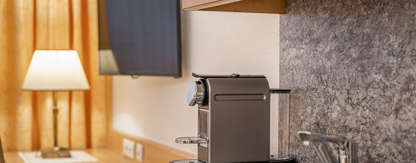 Eine moderne Küchenarbeitsplatte mit einem Spülbecken aus Edelstahl, einer eleganten Kaffeemaschine und einem Wasserhahn. Im Hintergrund befinden sich ein Flachbildfernseher und eine Tischlampe neben gelben Vorhängen.