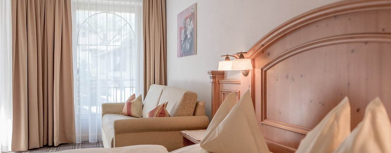 Ein gemütliches Apartmentzimmer mit einem hölzernen Kopfteil, ordentlich angeordneten cremefarbenen Kissen und Bettzeug, einem beigen Sofa mit Akzentkissen, großen Fenstern mit durchsichtigen Vorhängen und einem gerahmten Kunstwerk an der Wand.