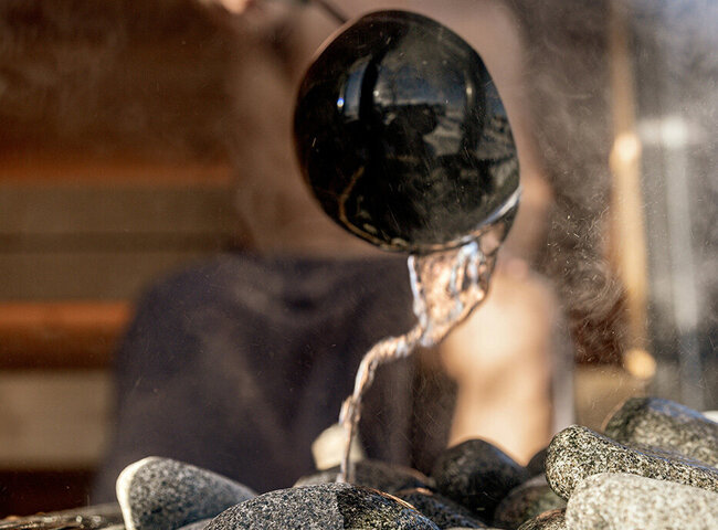 Eine Person gießt Wasser aus einer Schöpfkelle auf heiße Saunasteine, wodurch Dampf entsteht. Das Bild ist auf die Kelle und die Steine fokussiert, während die Person im Hintergrund verschwommen ist.