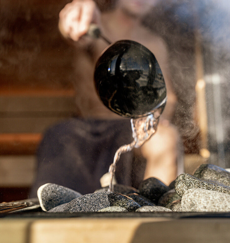 Eine Person gießt Wasser aus einer Schöpfkelle auf heiße Saunasteine, wodurch Dampf entsteht. Das Bild ist auf die Kelle und die Steine fokussiert, während die Person im Hintergrund verschwommen ist.