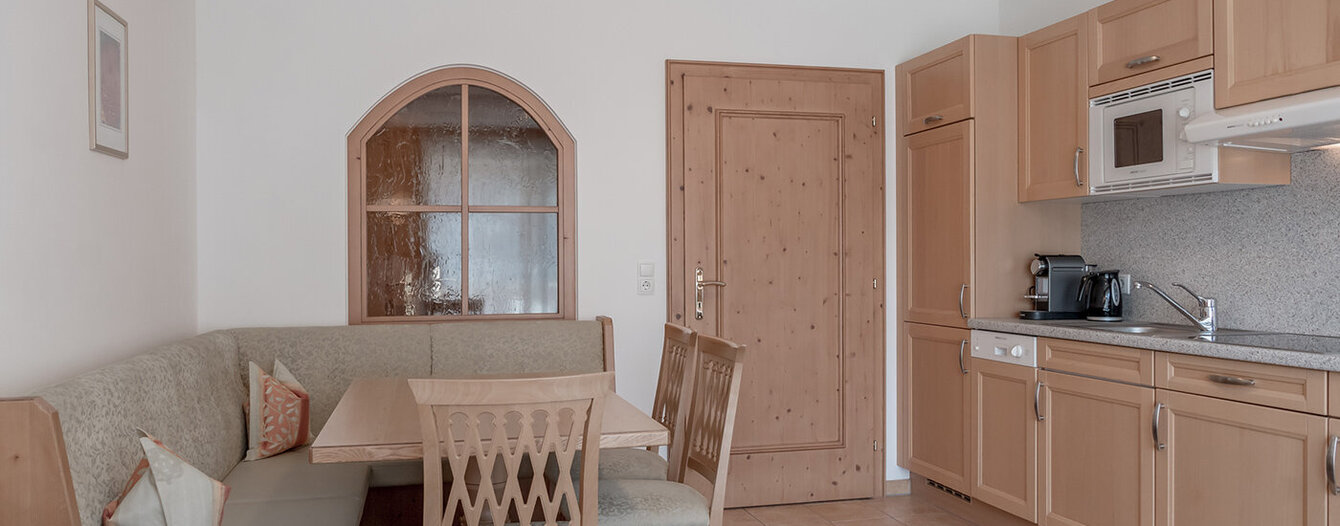 Eine gemütliche Küche mit hellen Holzschränken, einer Eckbank mit beiger Polsterung, einem hölzernen Esstisch mit Stühlen, einer Mikrowelle, einer Kaffeemaschine, einem Geschirrspüler sowie einer Tür und einem Rundbogenfenster im Hintergrund.