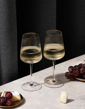 Zwei Gläser mit Weißwein auf einer hellen Fläche, daneben rote Weintrauben und Weichkäsestücke auf kleinen Tellern; im Hintergrund dunkle Vorhänge.