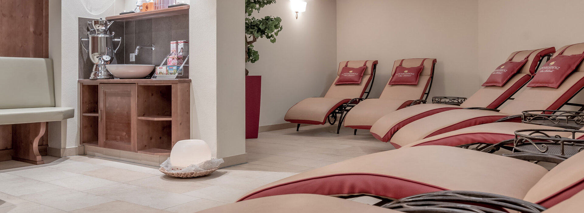 Eine entspannender Wellnessbereich mit gepolsterten Liegen, einem Holzregal und darüber die Teebar. Die Atmosphäre ist ruhig und einladend.